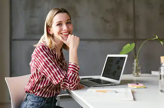 Person sitting at a desk with a laptop, papers, and a small plant, facing the camera.