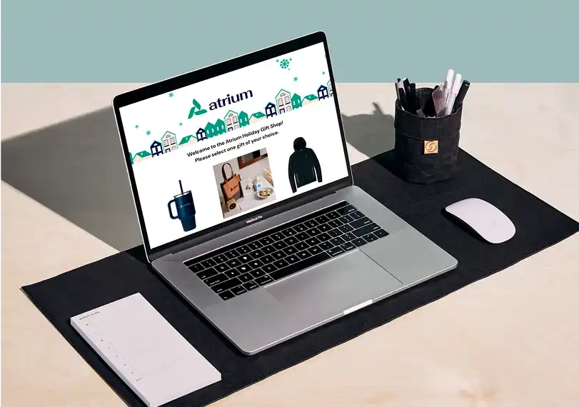 Online company gift shop displayed on a laptop on a desk with mouse, pen holder, and notepad.