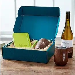 Open gift box with a wine bottle, mug, packaged treats, and shredded paper filler on a table.