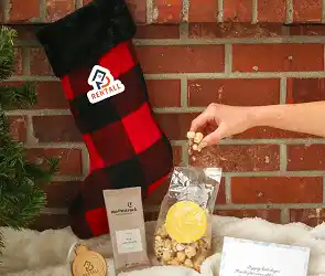 Holiday-themed gift set with a red and black plaid stocking, packaged snacks, and a hand reaching into a clear bag.