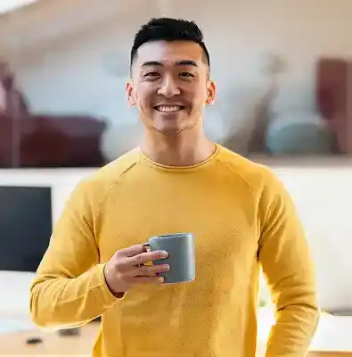 Person holding a mug while standing in a home office with a desk and computer in the background.